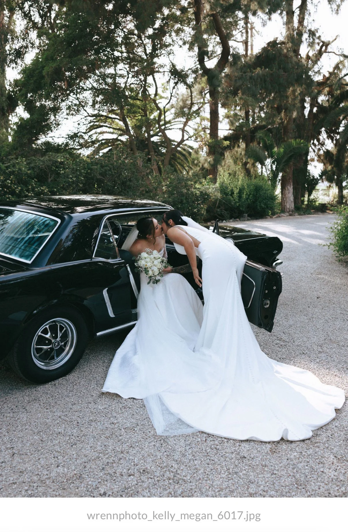 Two brides in wedding dresses sharing a kiss outside a black vintage car, surrounded by trees.
