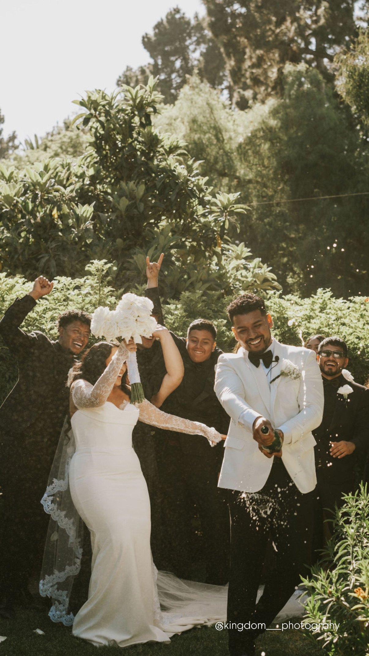 A wedding celebration outdoors with a bride in a white gown and veil, holding a bouquet, and a groom in a white tuxedo and bow tie, opening a champagne bottle. Several guests are around them, cheering and throwing confetti, with lush greenery and tre