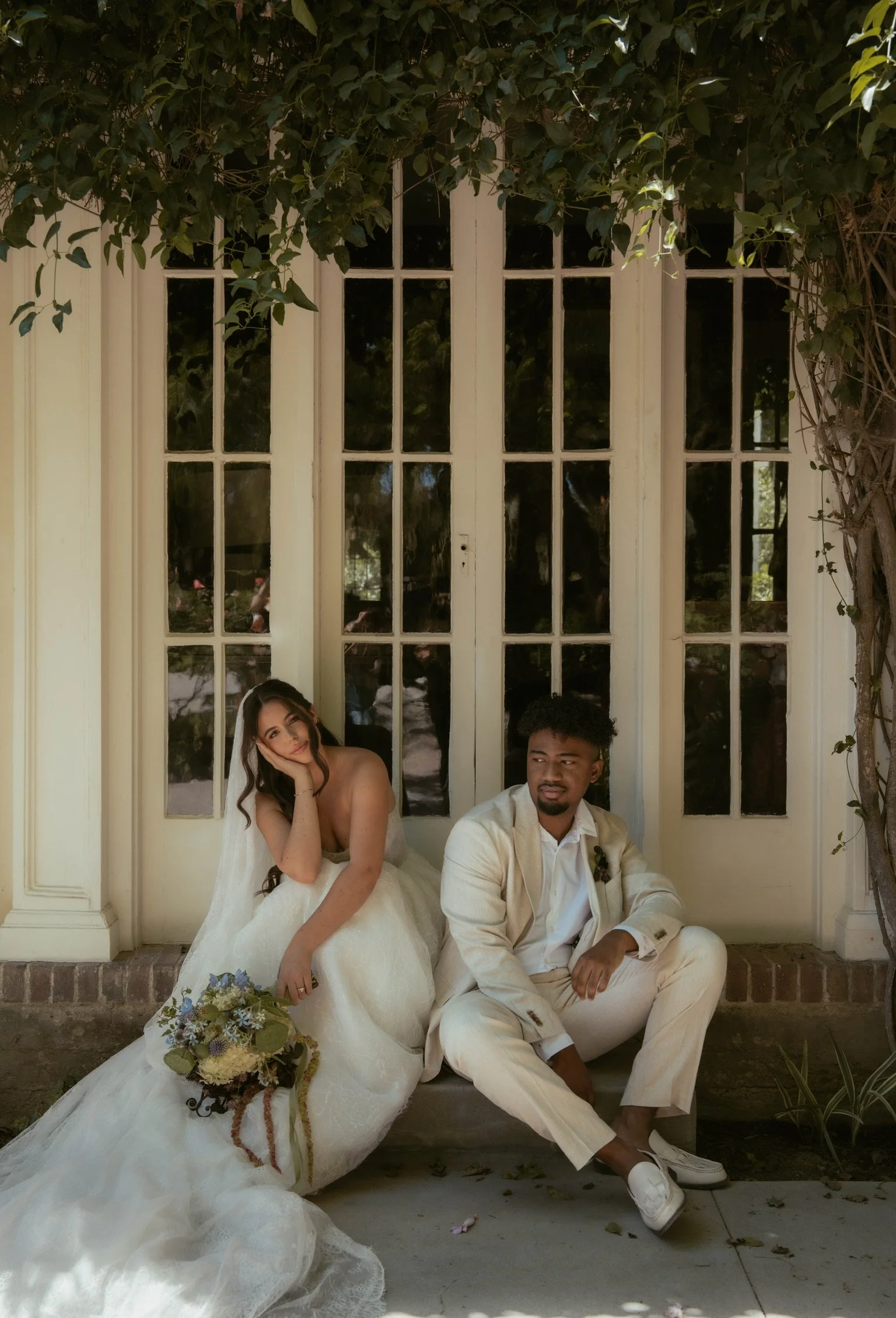 A bride and groom sitting side by side outside a building with large windowed doors. The bride is wearing a white wedding dress and holding a bouquet, while the groom is in a light-colored suit with a white shirt and white shoes. The woman rests her 