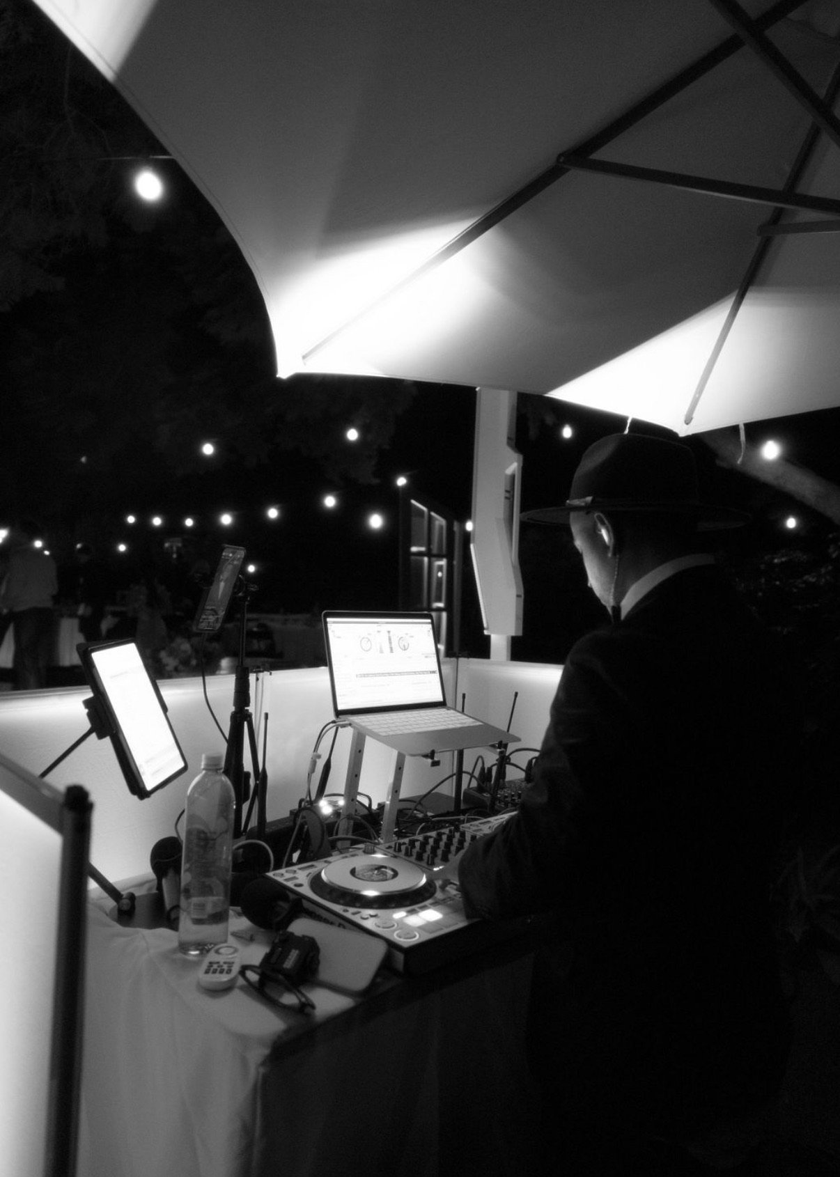 A DJ wearing a hat and glasses is performing at an outdoor event at night, surrounded by equipment, under a large umbrella with string lights overhead.