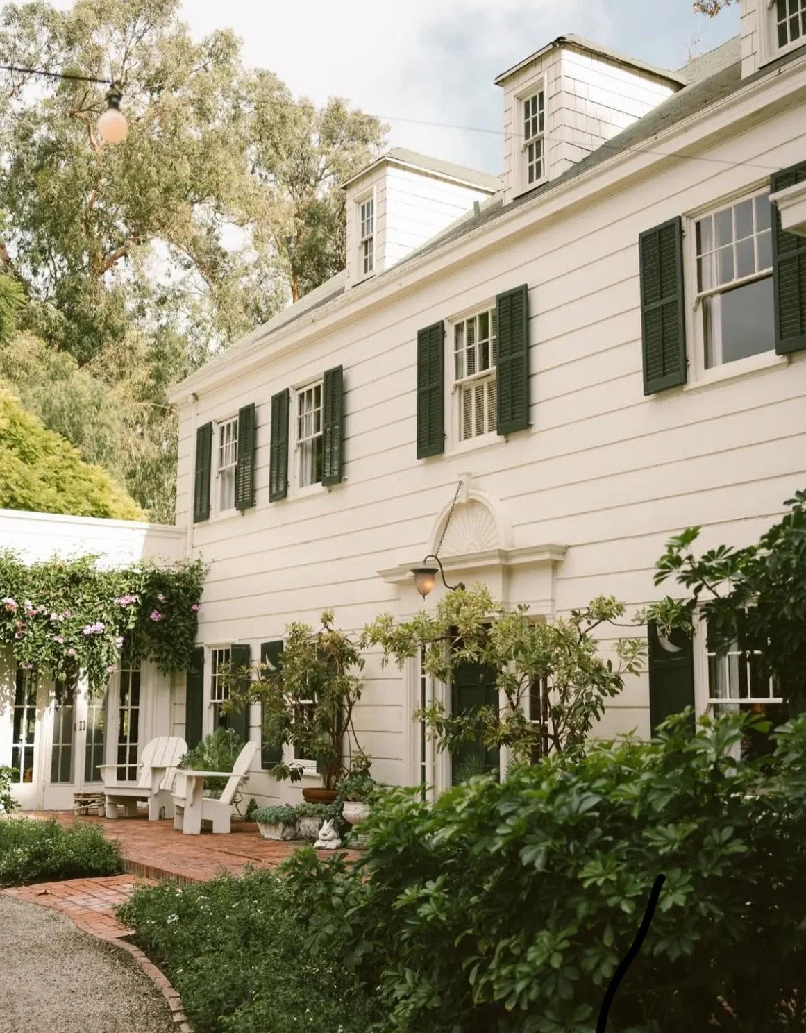 White two-story house with black shutters, a brick porch, and surrounding greenery and trees.