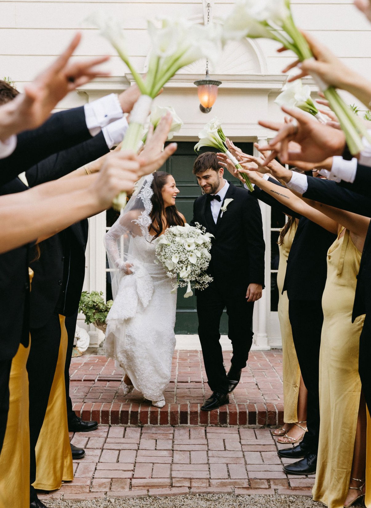 Bride and groom walking out of a building surrounded by friends tossing white calla lilies in celebration.