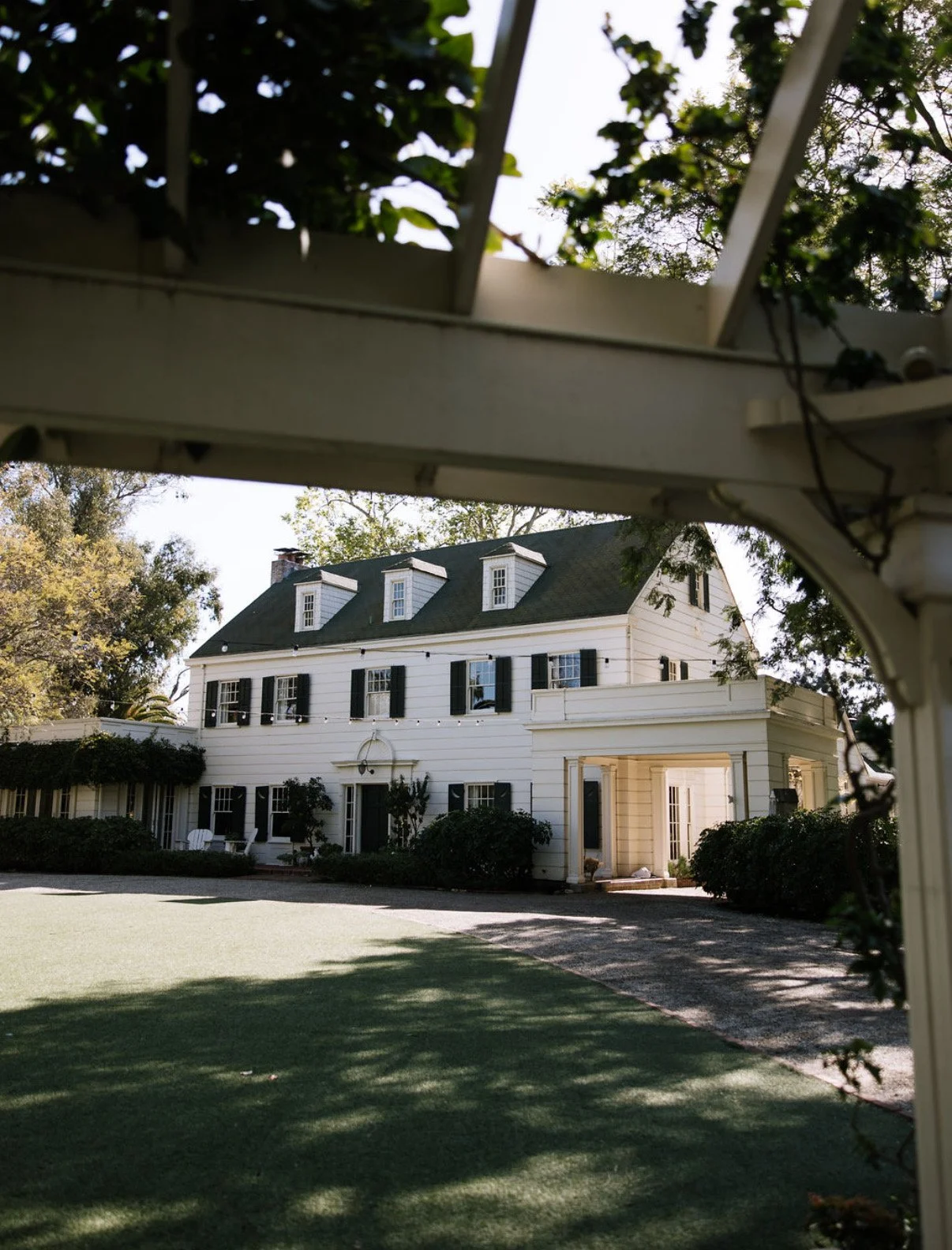 View of a large, white, three-story house with black shutters, dormer windows on the roof, and a small overhang porch at the front door, seen through a decorative white trellis with vines and greenery in the foreground.