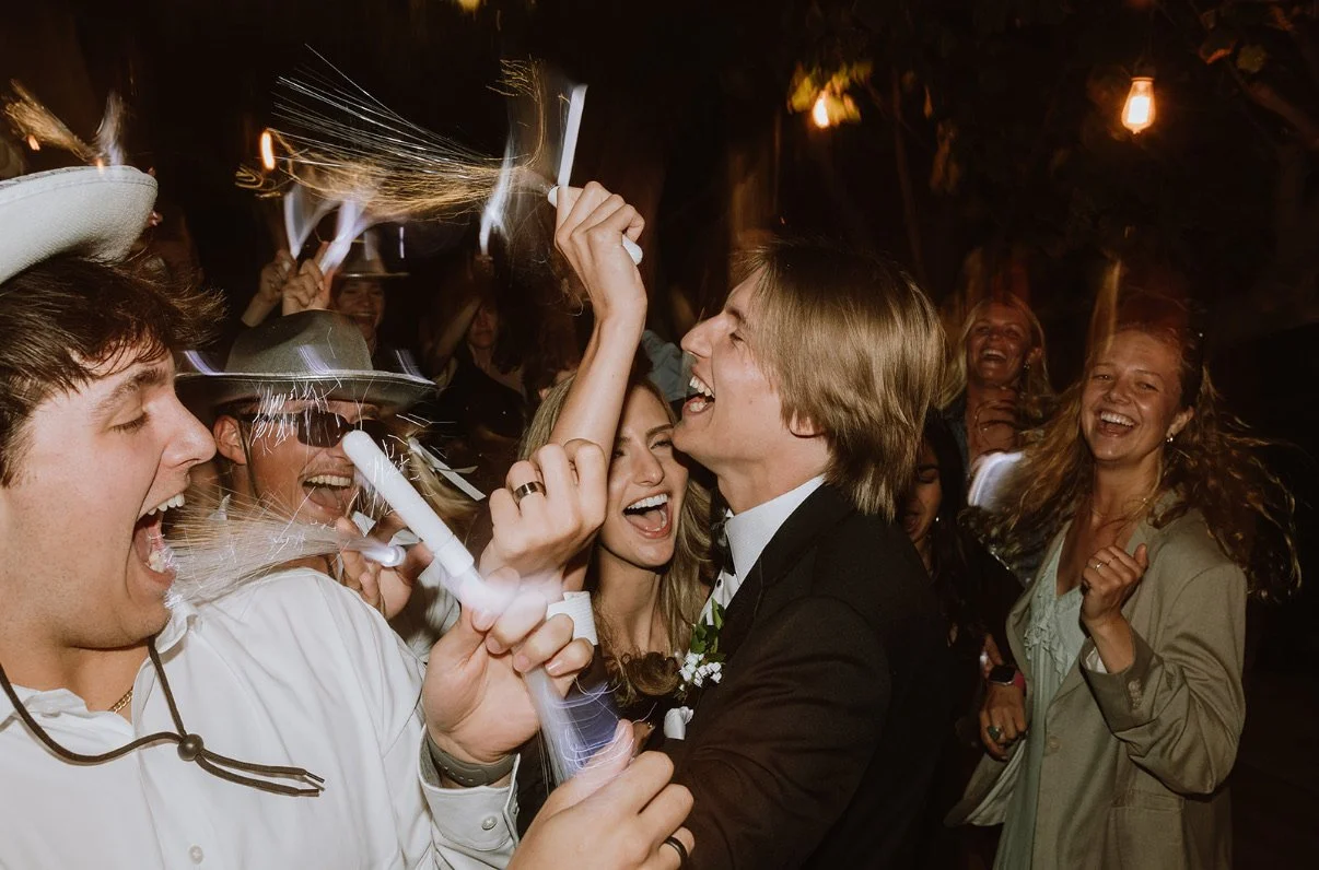 People celebrating at night, some wearing hats and holding sparklers, smiling and laughing.