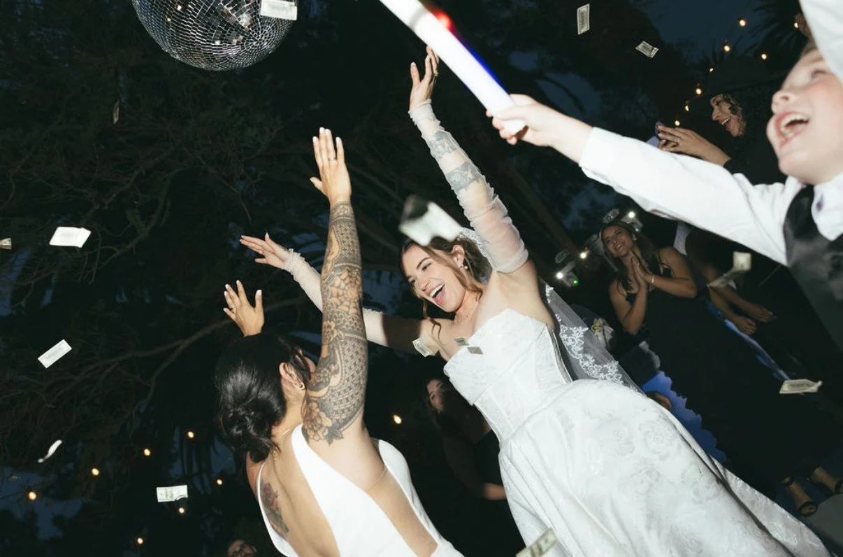 Two brides dance with their families with a disco ball twinkling above them at their reception on the lawn at the McCormick Home Ranch.