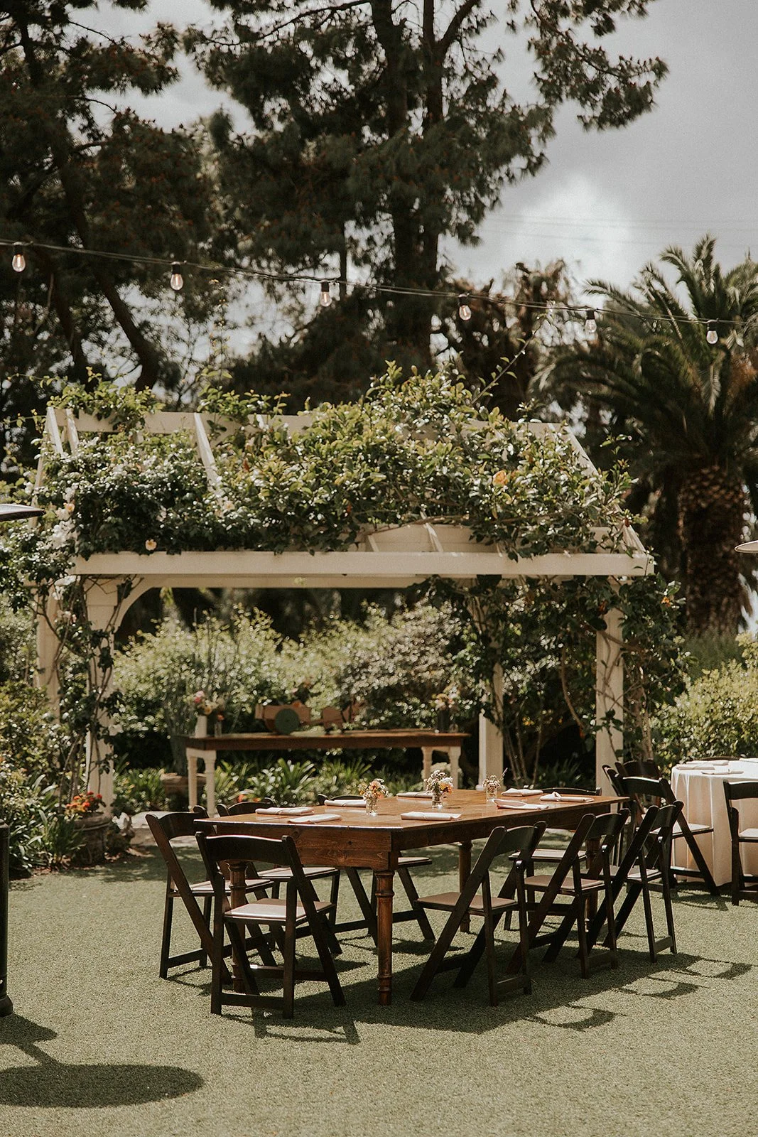 Outdoor wedding reception area with a wooden table and black chairs on artificial grass, decorated with small floral arrangements, surrounded by lush greenery and white floral arch, with string lights hanging from the trees.
