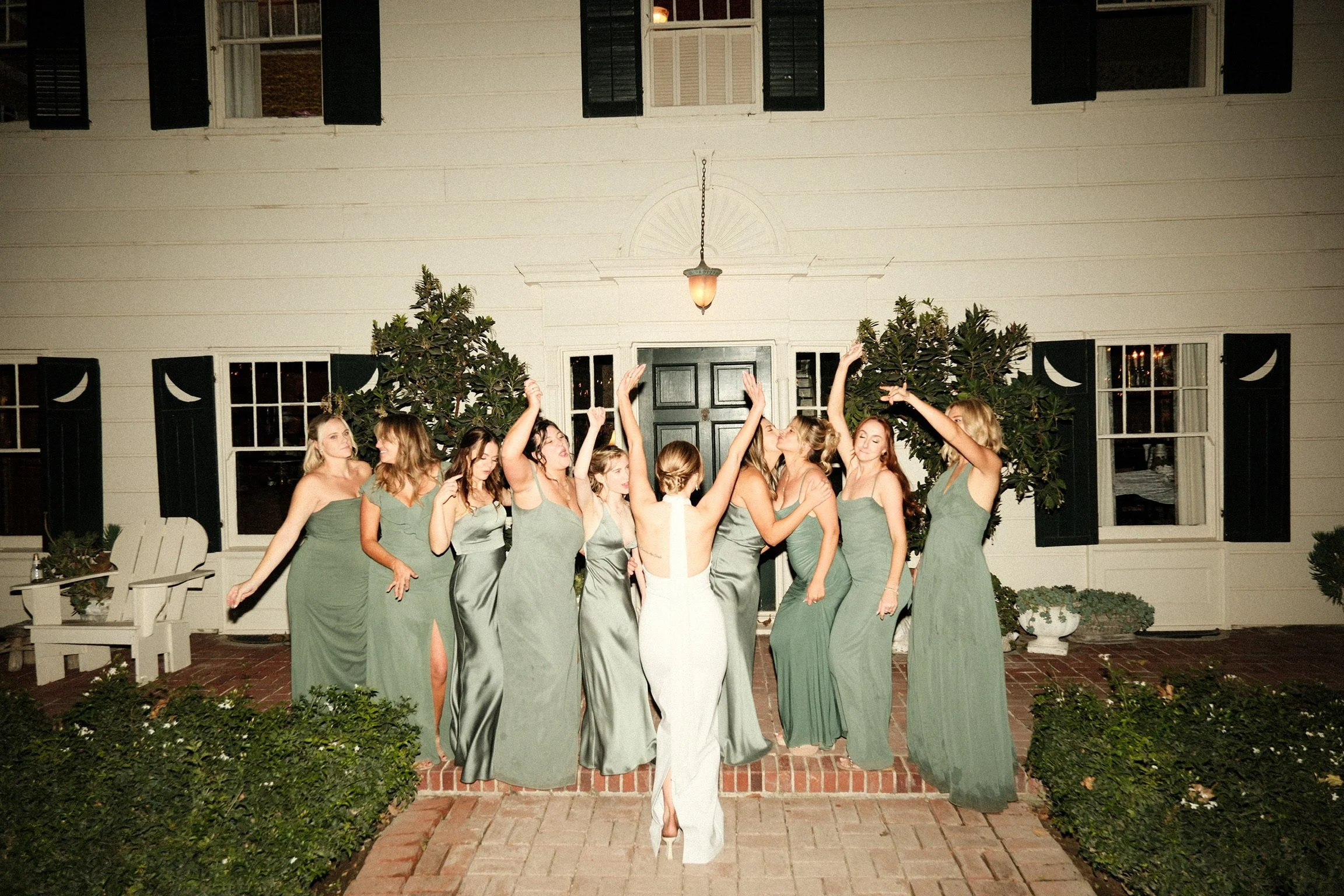 Bride wearing backless satin dress dancing with hands up towards her bridesmaids dressed in green also dancing in front of McCormack Home Ranch