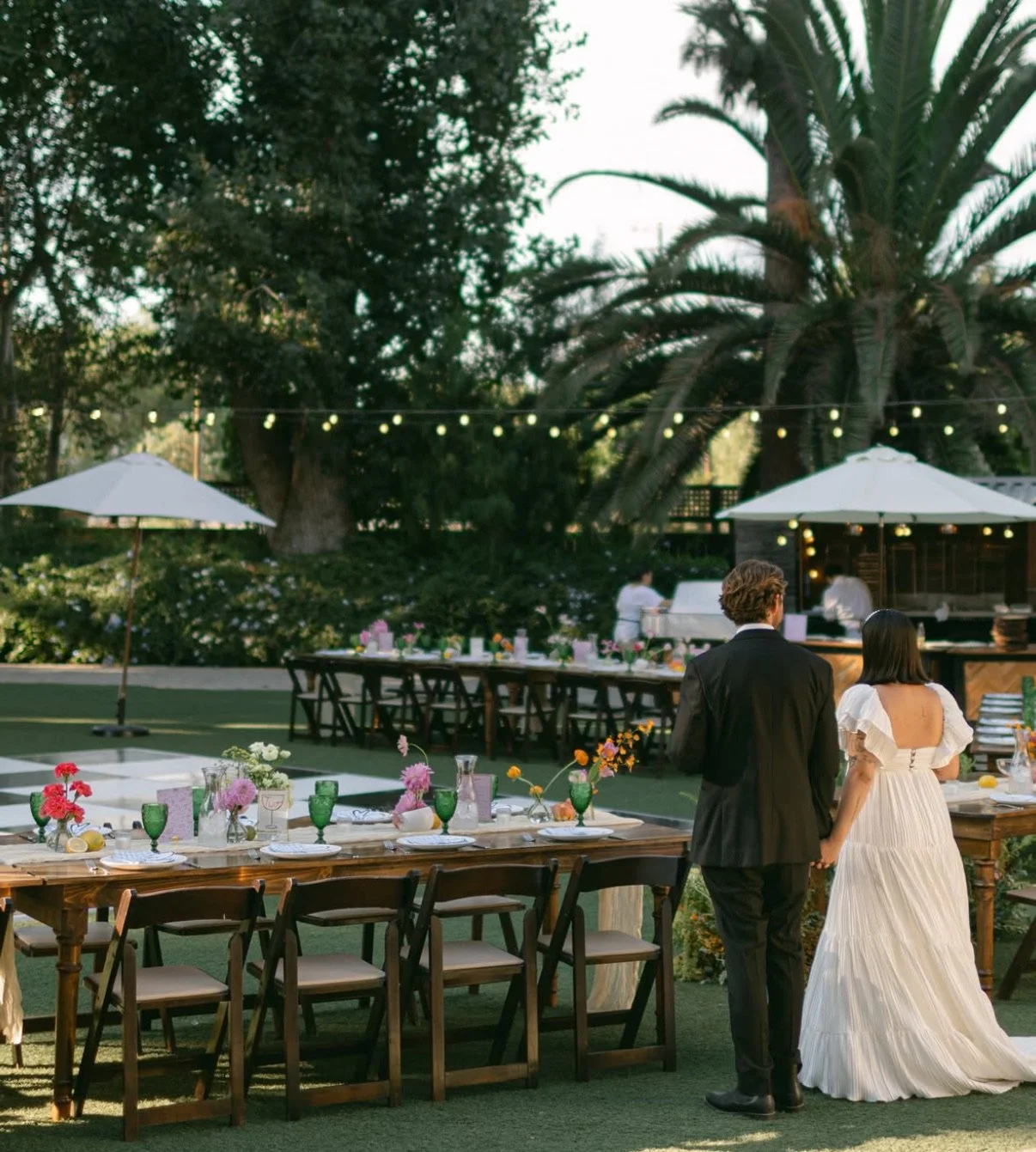 Bridal couple looks out on the color table settings for their evening wedding event. Catering can be seen in background with fairy lights twinkling.