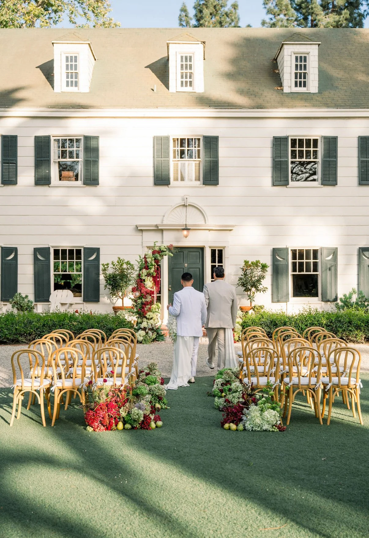 Two grooms walking towards an outdoor wedding ceremony setup in front of a white house with dark green shutters.