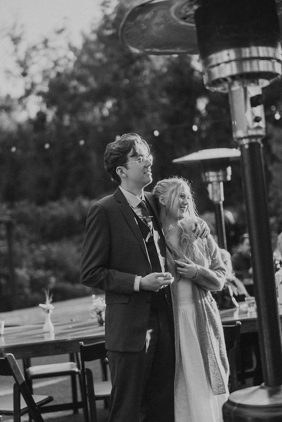 A young couple stands outdoors at a gathering, smiling and embracing. The man wears glasses and a suit, and the woman has long, wavy hair and wears a cardigan over a dress.