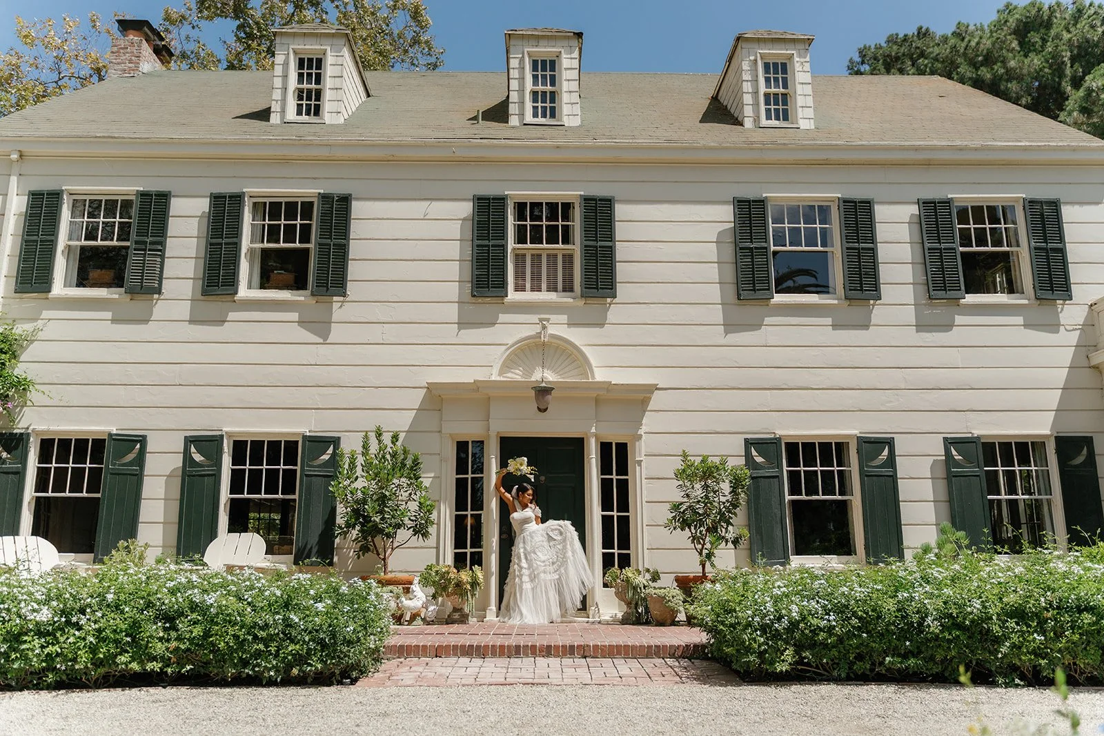 A woman in a wedding dress standing on the front steps of a white colonial-style house with green shutters, holding a bouquet and lifting her dress.