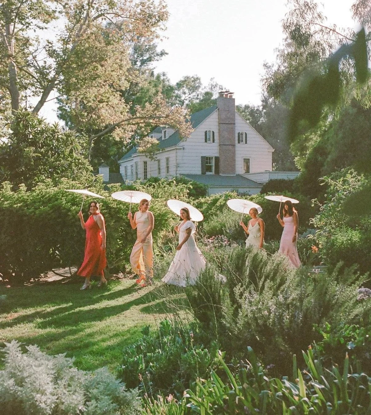 Bridal party walking out to garden holding paper umbrellas wearing spring colored dresses.