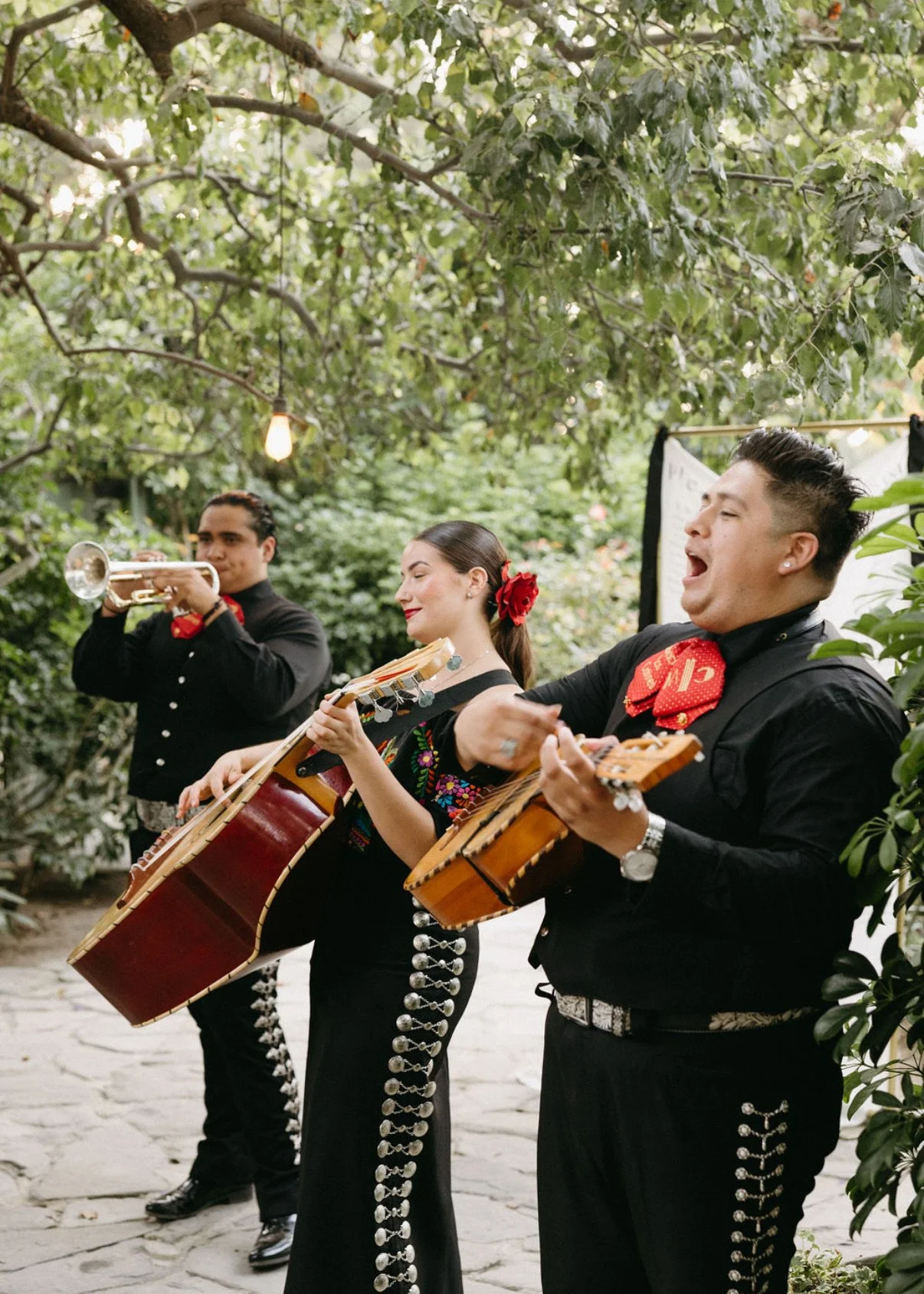 Three musicians performing outdoors, dressed in traditional Mexican attire. Two are playing guitars, and one is playing a trumpet. The women wear black dresses with colorful embroidery and red flowers in their hair. The men wear black shirts with sil