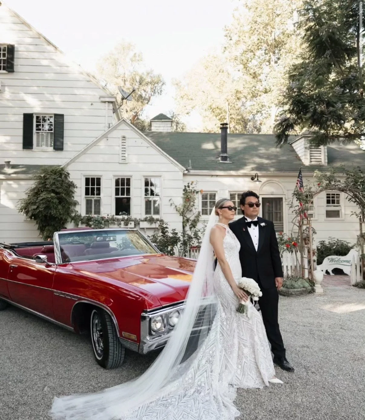 A bride and groom in wedding attire standing next to a red vintage convertible car outside a white house with greenery and decorative flowers.