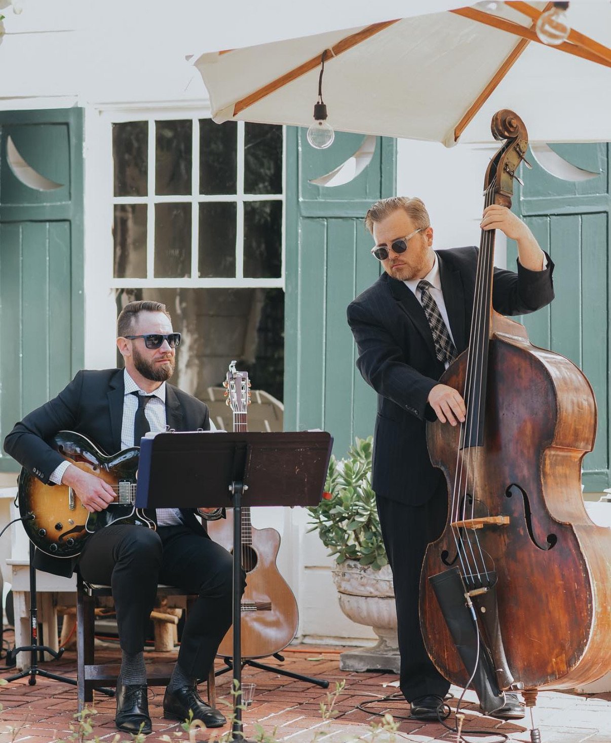 Two musicians, both in suits and sunglasses, performing outdoors. One is seated with a guitar, and the other is standing, playing a double bass, under a large umbrella.