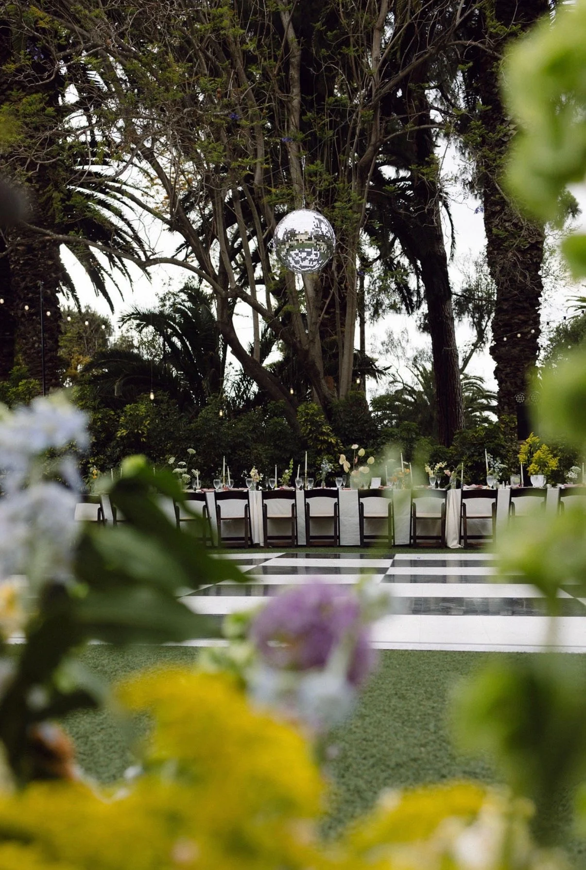 Outdoor event setup with long dining table, chairs, and floral centerpieces, surrounded by greenery and trees, with a disco ball hanging from tree branches.