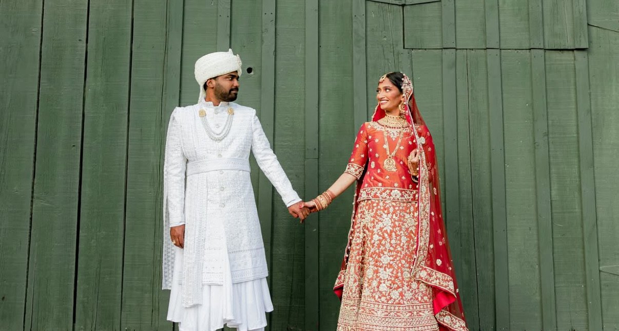 A bride and groom in traditional Indian wedding attire holding hands, standing in front of a green wooden wall.