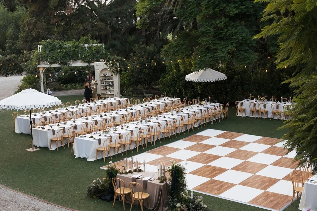 Outdoor wedding reception setup with long dining tables, white tablecloths, wooden chairs, a checkered dance floor, and string lights in a garden setting.