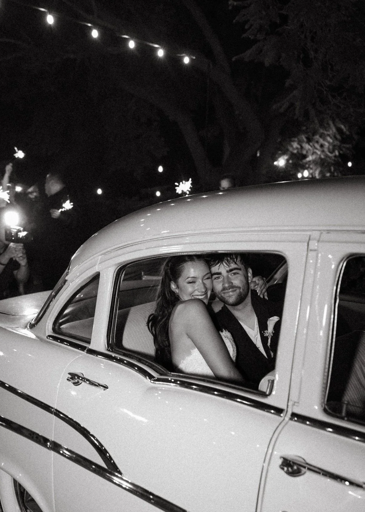 Black and white photo of smiling bride and groom in vintage wedding car with sparklers in background
