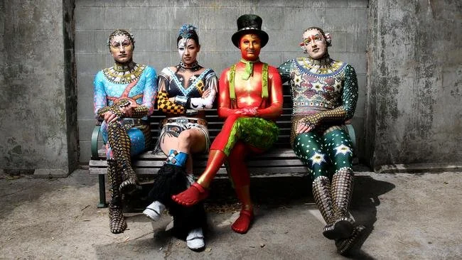 Four women wearing colorful, eccentric costumes sitting on a bench against a concrete wall, with unique makeup and poise.