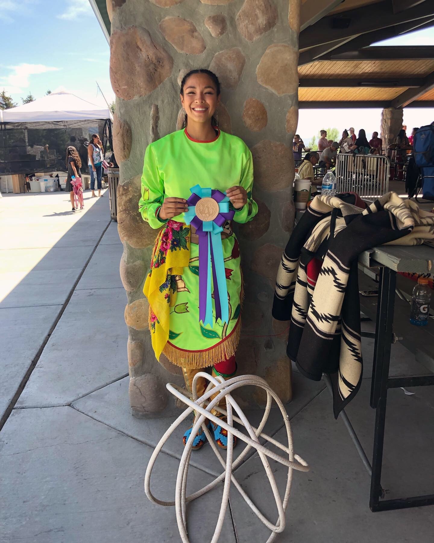 Young woman smiling and holding a blue and purple ribbon with a gold medal, standing against a stone pillar, wearing a bright green shirt and a colorful skirt, with a wheelchair in front of her and a group of people in the background.