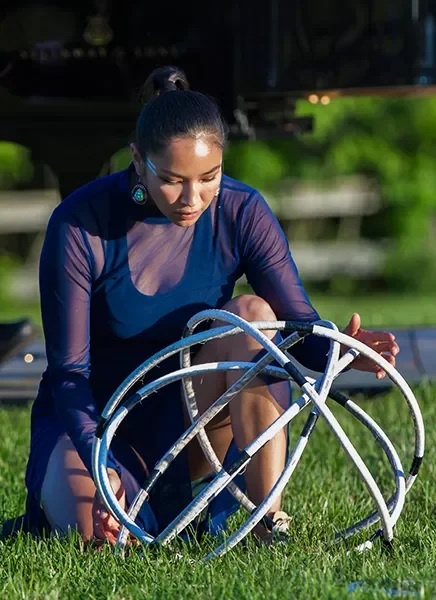A woman in a blue dress is kneeling on green grass, looking down at a metallic sculpture or art piece with curved white tubes.