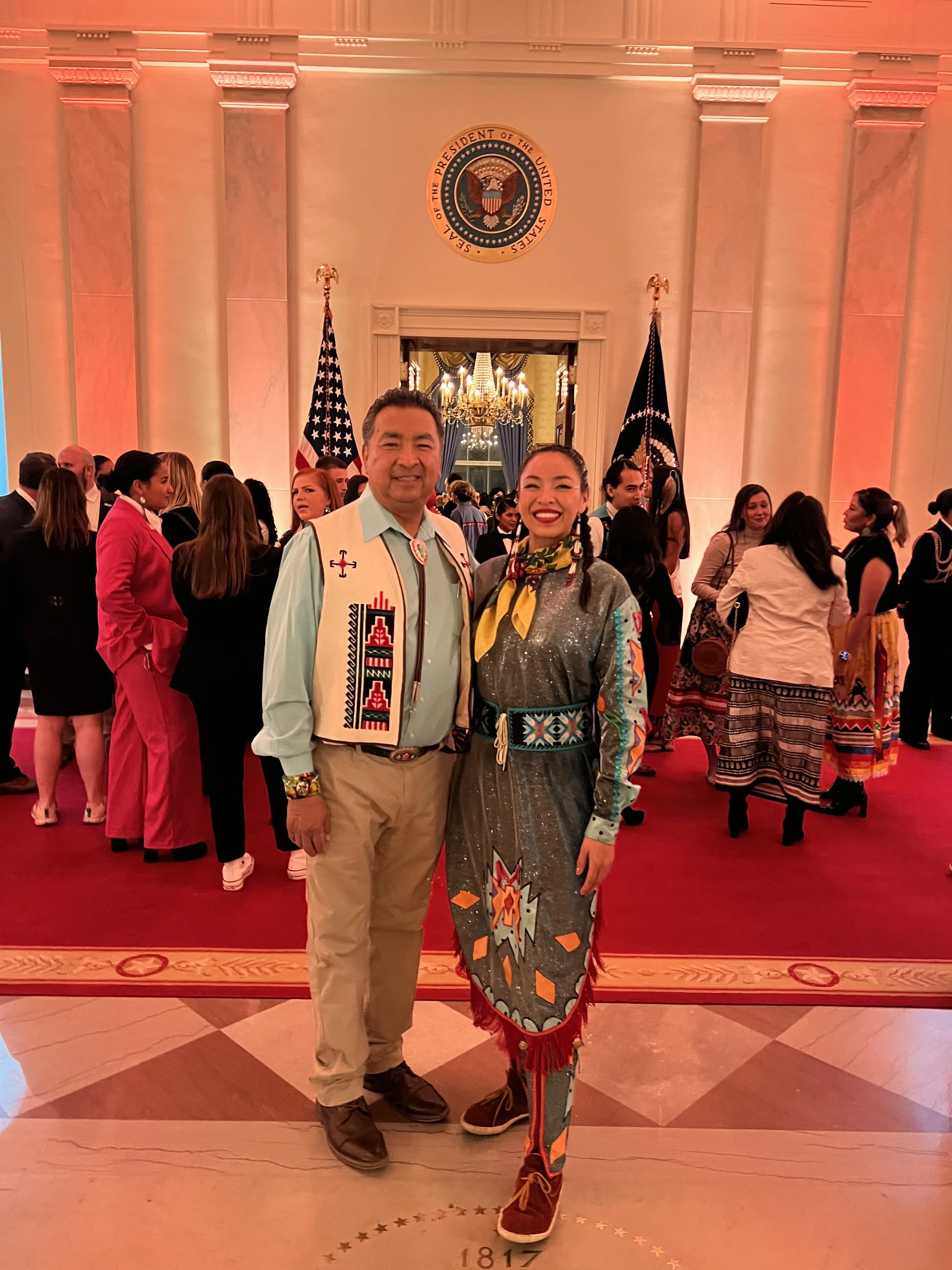 Two people smiling and standing together inside the White House, with a group of people in the background. The man is wearing traditional indigenous attire, and the woman is dressed in colorful clothing. The White House seal is visible above them.