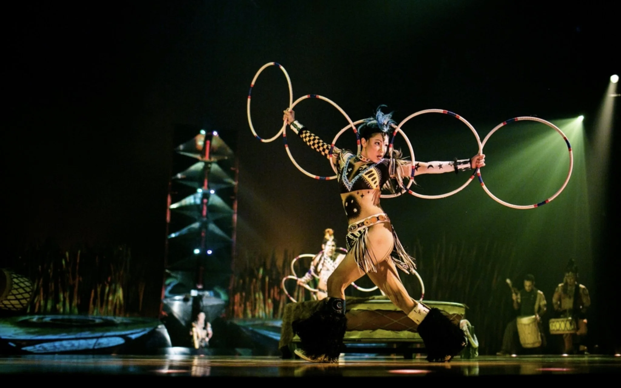 Performer in colorful costume spinning hoops on stage with stage lights and other performers in background.