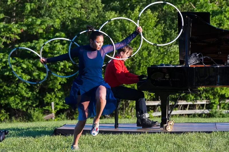 A woman and a man performing outdoor acts, with the woman hula hooping and the man playing a grand piano on a grassy field with trees in the background.