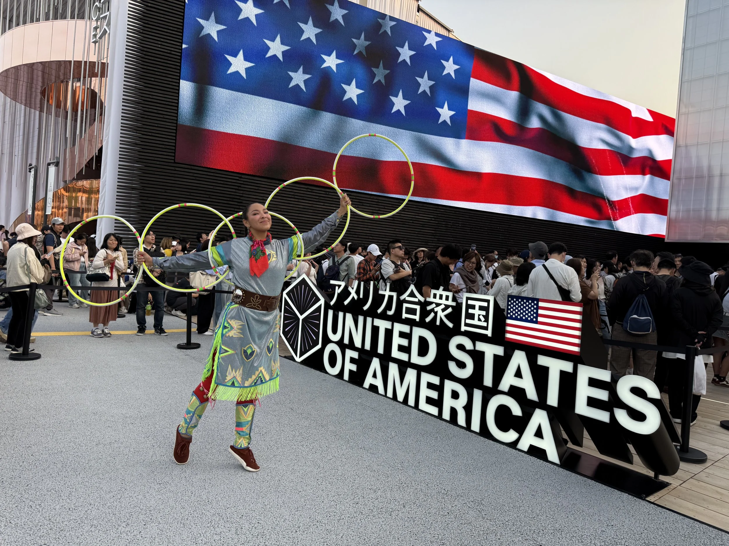 Woman in colorful traditional Native American attire performing with hoops at a celebration in front of a sign that reads 'United States of America' with an American flag, and a large American flag displayed on a building.