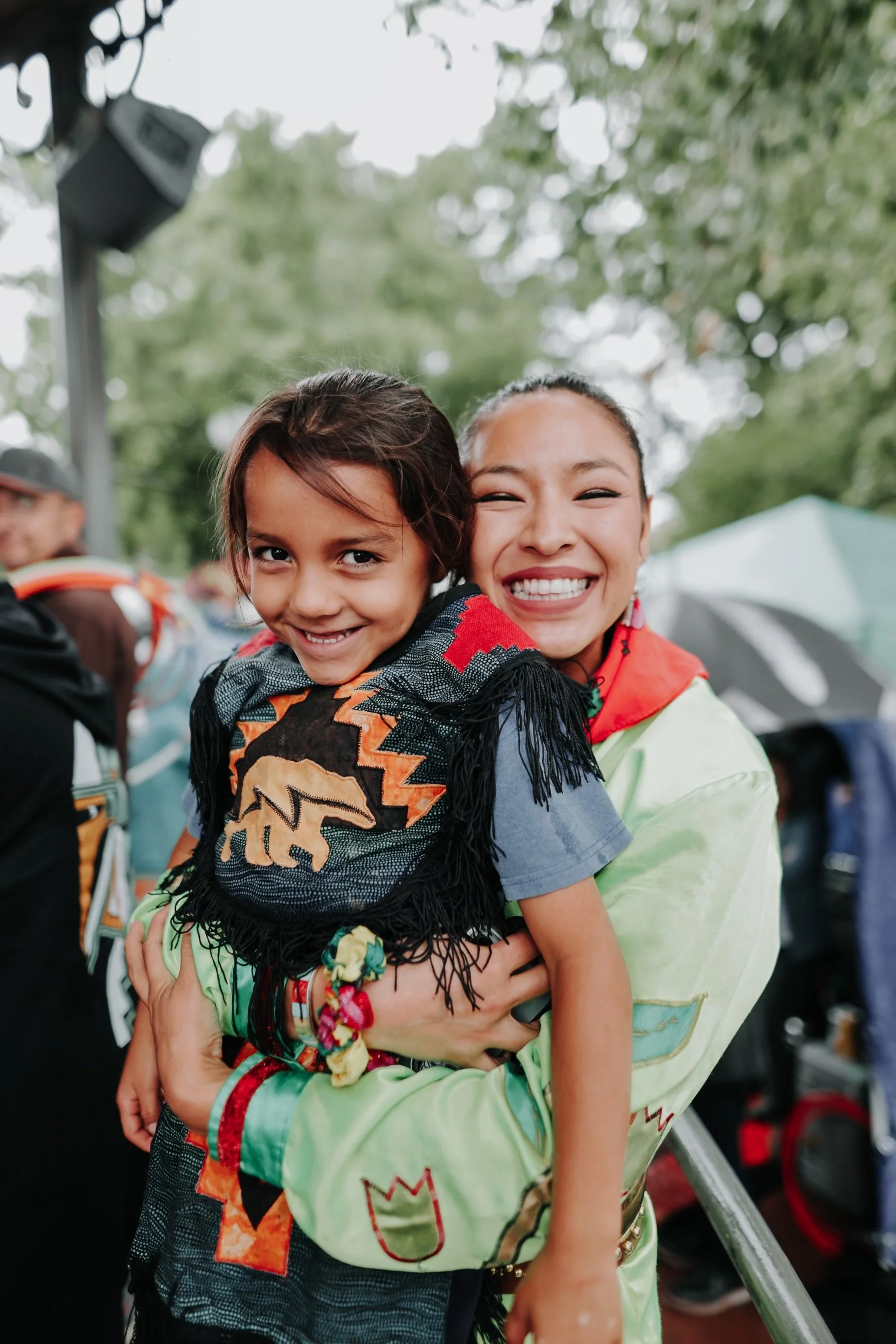 A woman and a young girl hugging outdoors, smiling at the camera. The girl is wearing a traditional woven garment with a geometric pattern, and the woman is dressed in a light green jacket. The background shows trees and people, some holding umbrellas.
