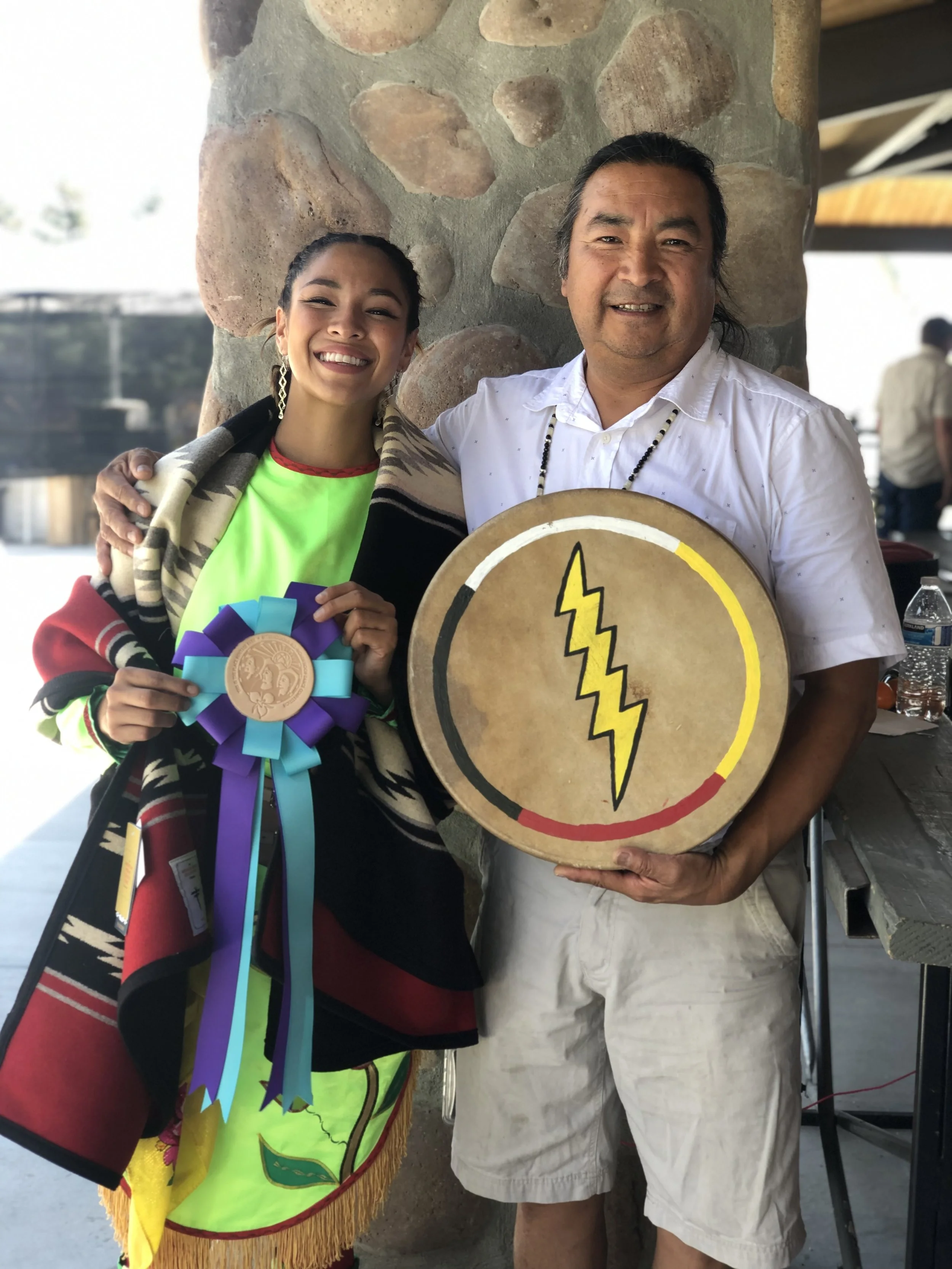 A young woman and an older man standing together, smiling. The woman is holding a large ribbon award. The man is holding a round sign with a lightning bolt symbol, and both are standing in front of a stone wall.