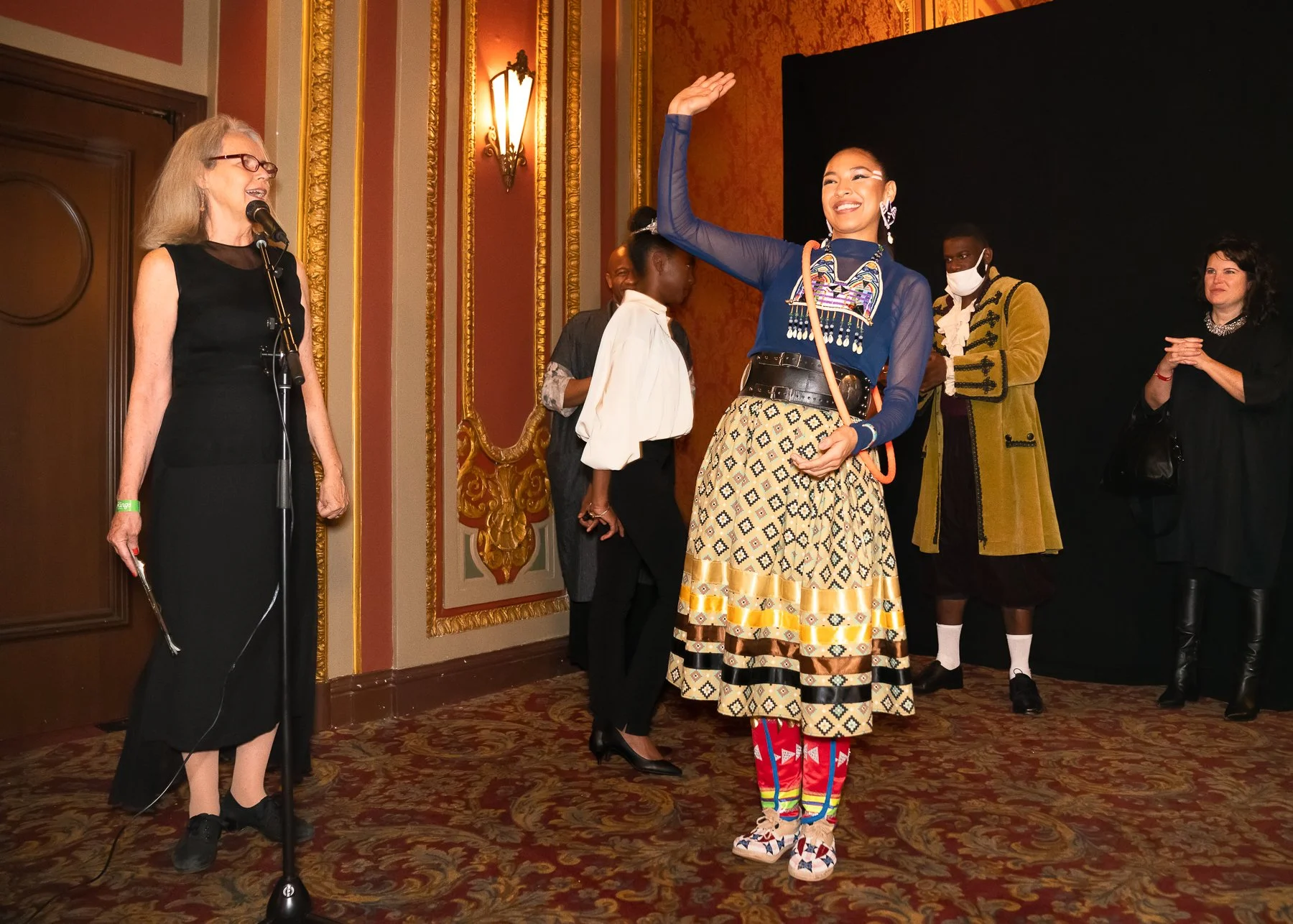 A group of women on stage at an event, with one woman in the center wearing vibrant traditional clothing, smiling and waving, while speaking into a microphone. Others stand around her, some clapping and smiling.