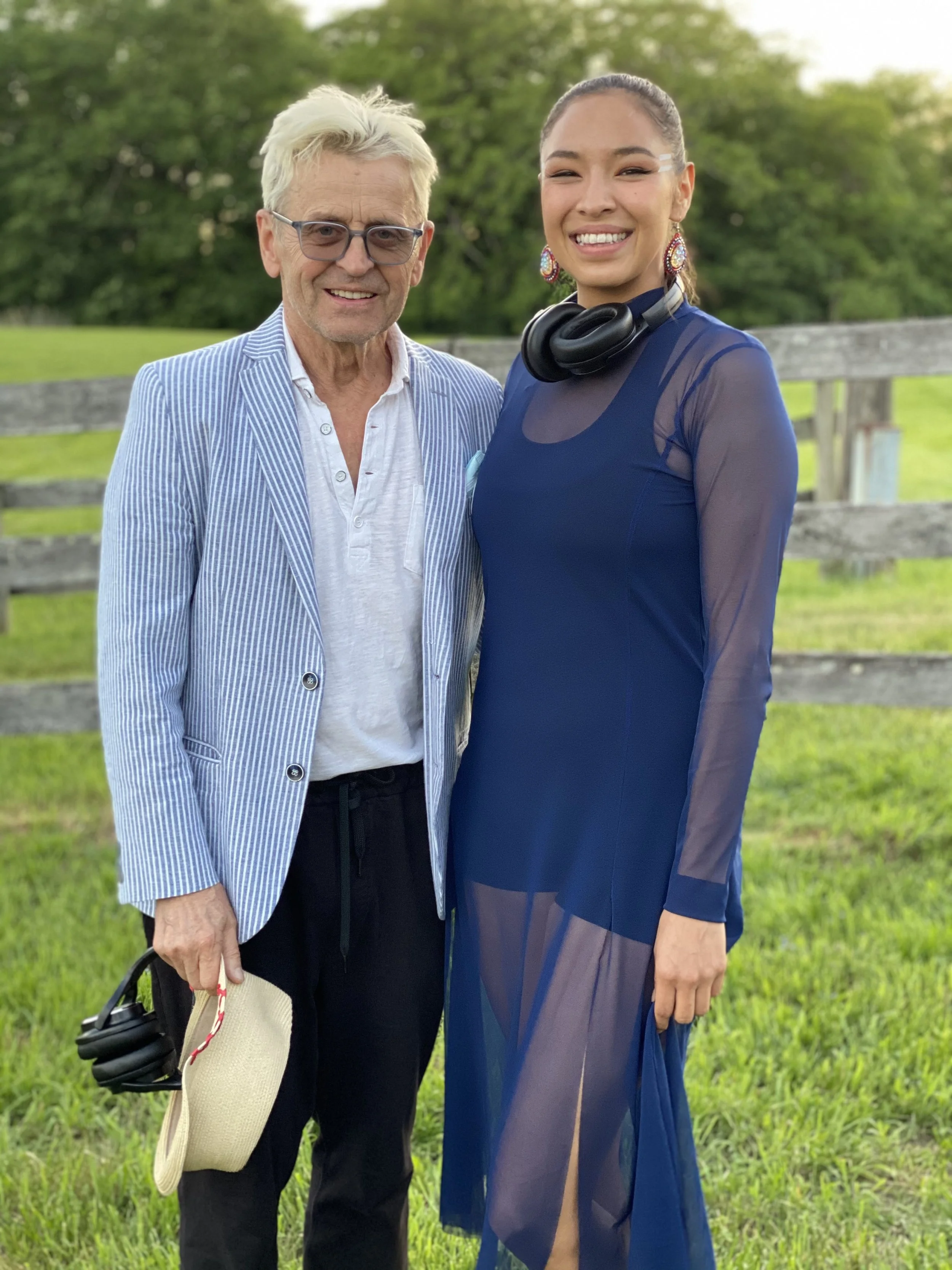 An elderly man and a young woman stand outdoors on grass, smiling at the camera. The man wears glasses, a light blue striped blazer, a white shirt, and black pants, holding a straw hat and headphones. The woman wears a sheer blue dress, colorful earr
