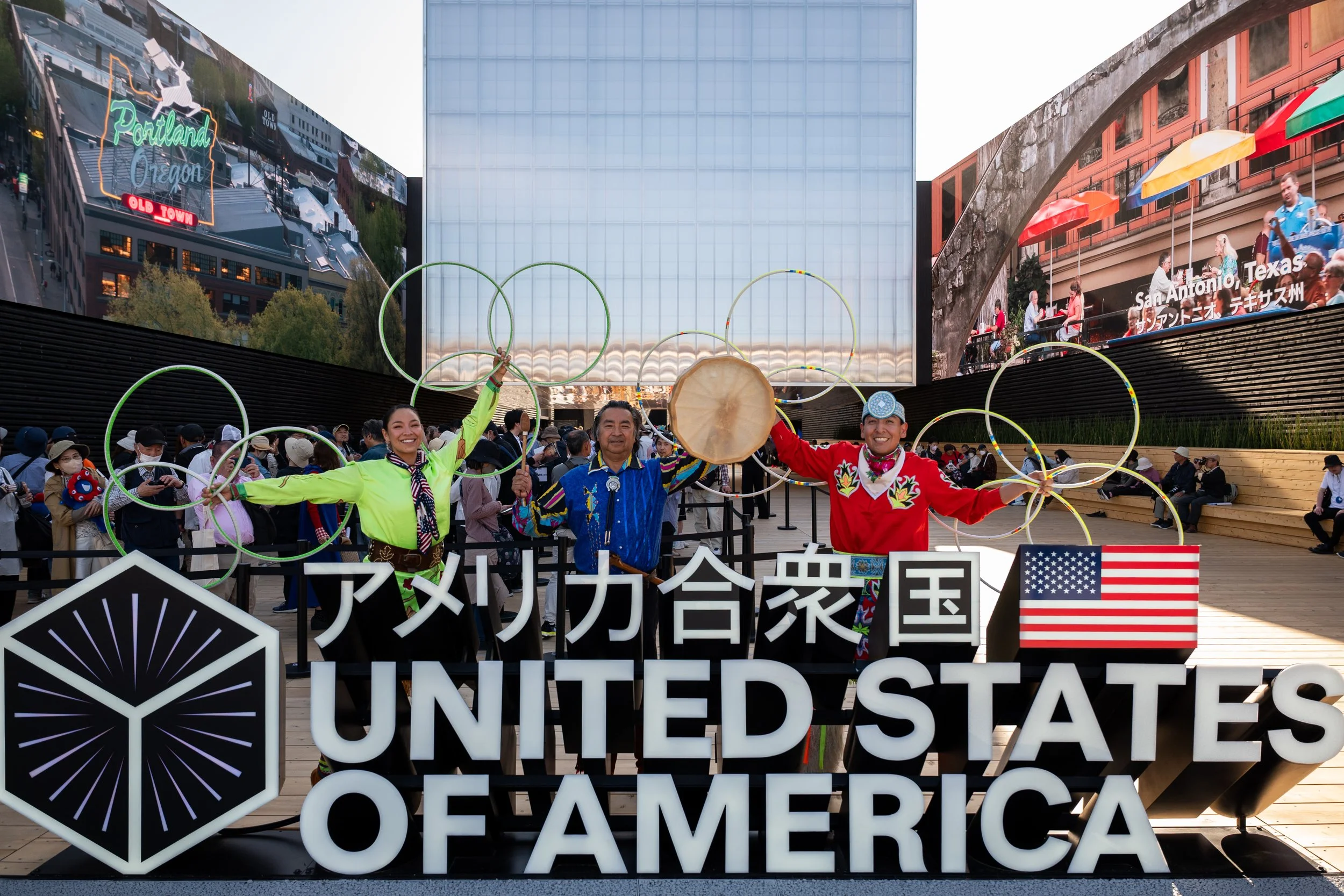 People celebrating with Olympic rings and a sign that reads "United States of America" in front of a modern building with digital screens, one showing Portland, Oregon, and another showing San Antonio, Texas.