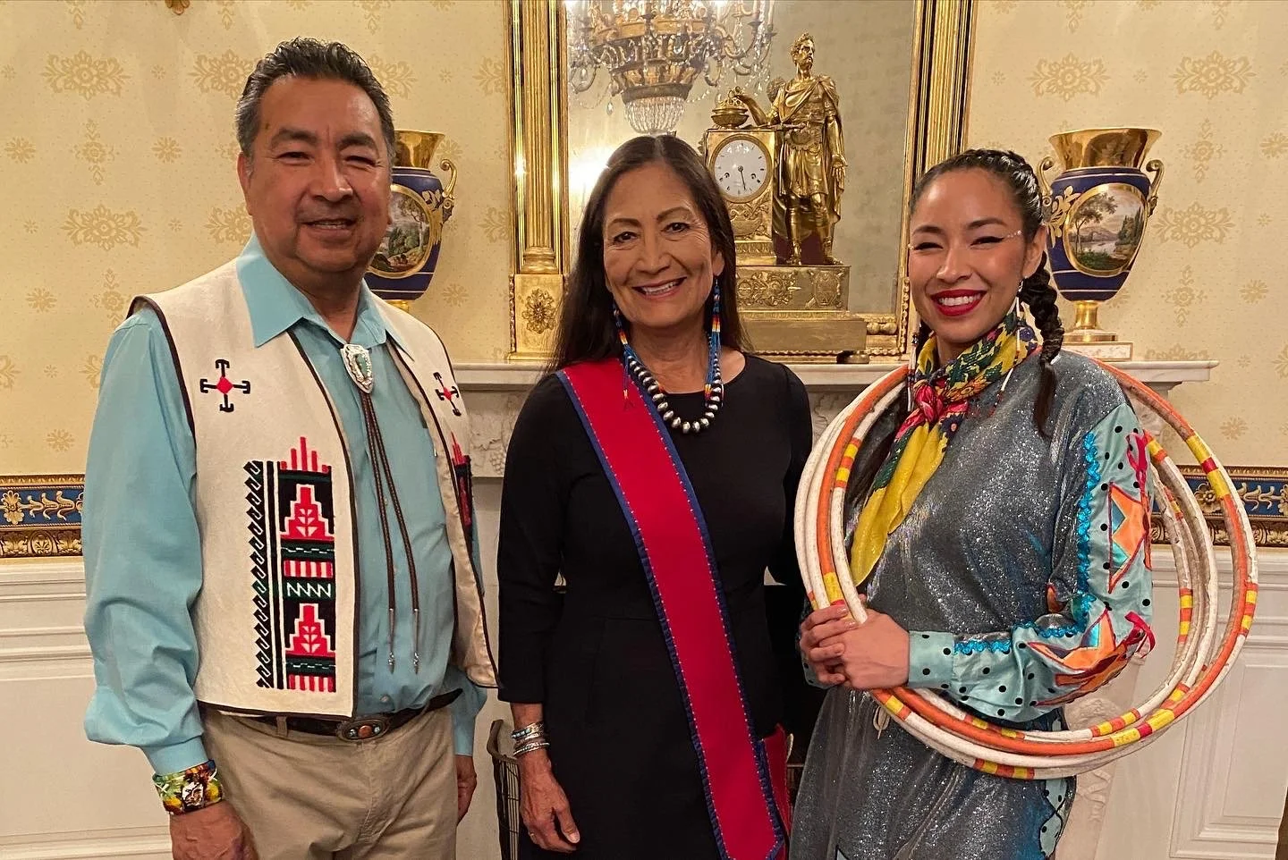 Three people standing in a decorated room, two women and one man. The man is wearing a light blue shirt with a white vest adorned with Native American-style patterns and jewelry. The woman in the middle is wearing a black dress with a red and blue sa