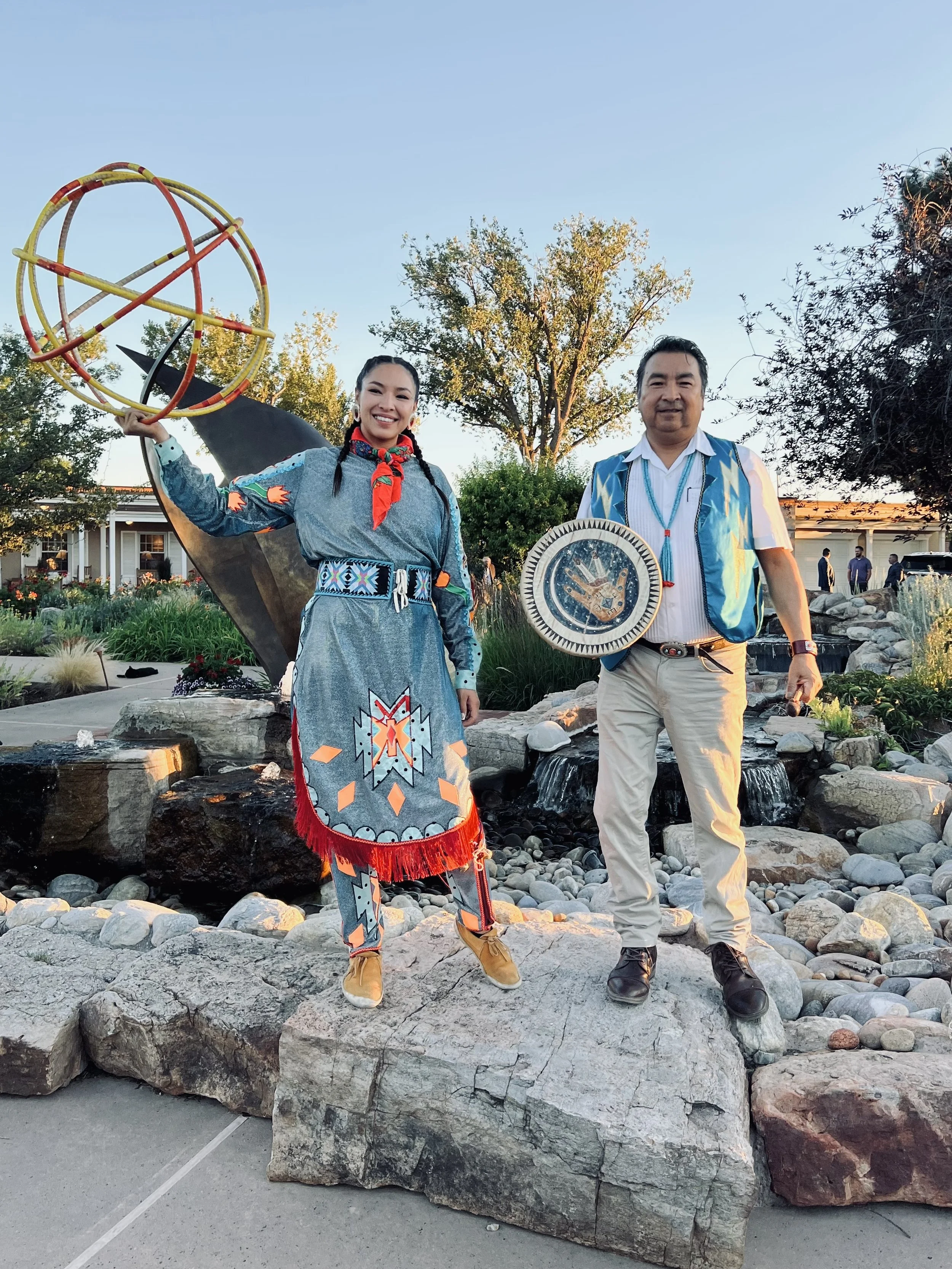 Two Native American individuals standing on rocks near a landscaped garden with trees and water features, dressed in traditional clothing, holding ceremonial items, with a blue sky in the background.