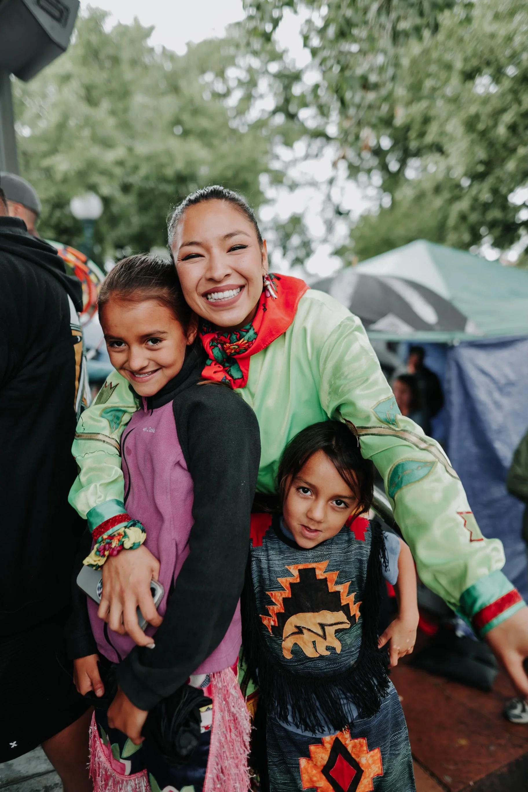 Smiling woman with long hair and earrings hugging two young girls, one in a pink and black jacket and another in traditional Native American clothing, at an outdoor event with tents and trees in the background.