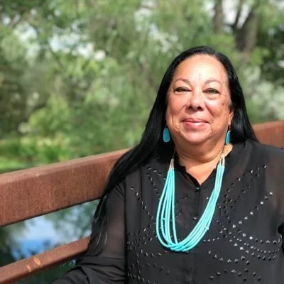 A smiling woman with long black hair wearing a black top and bright turquoise jewelry, standing outdoors with green trees in the background.