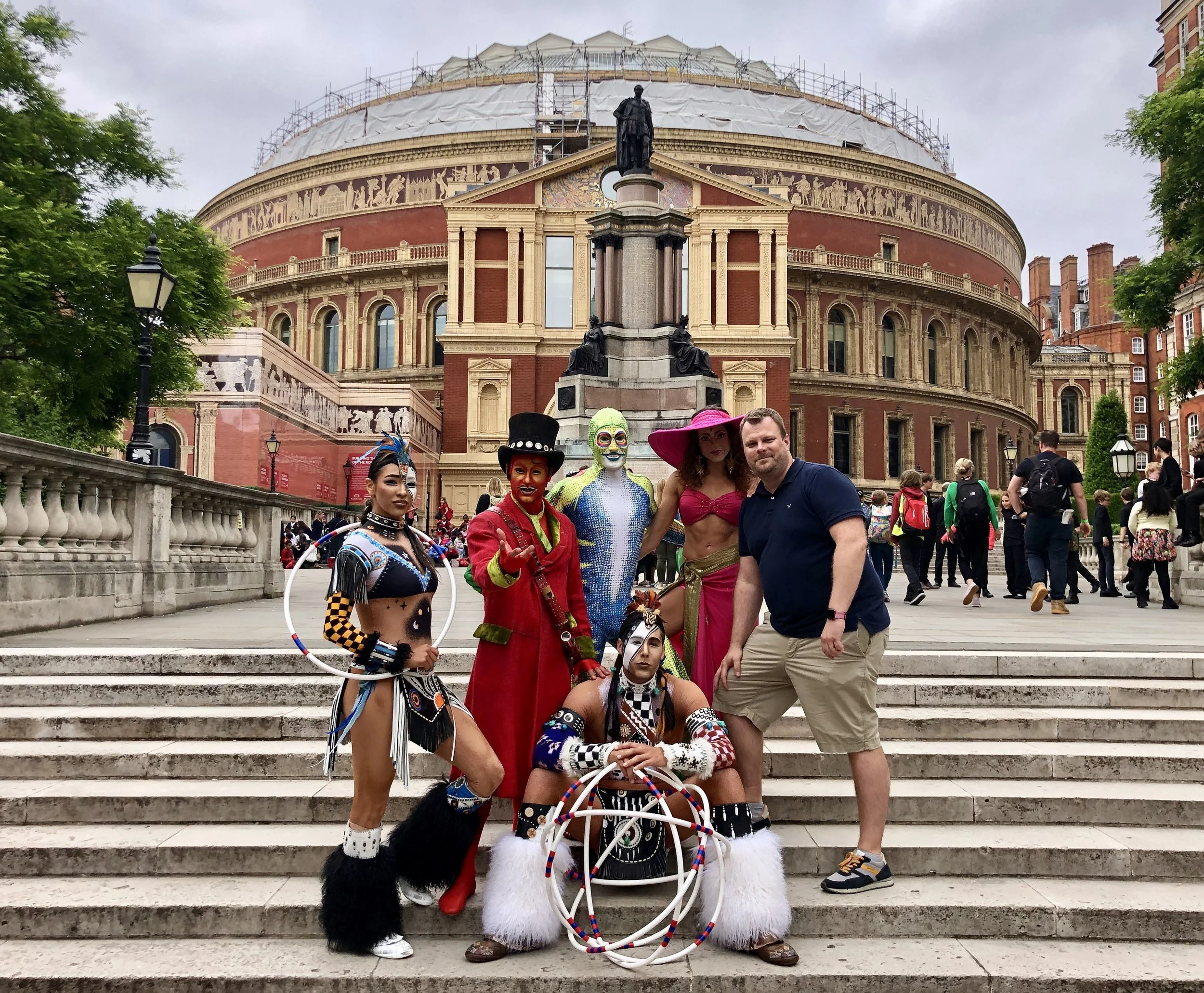 A group of five people in colorful costumes, including two women, two men, and a man dressed as a clown, posing on steps outside a historic theater or opera house with a large dome and statues, surrounded by other visitors.