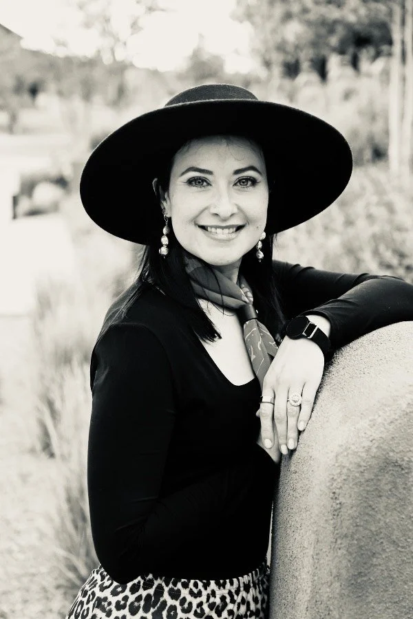 A woman smiling outdoors, wearing a large black hat, earrings, a scarf, a watch, a ring, and a black long-sleeve top, leaning on a stone structure with a natural background.