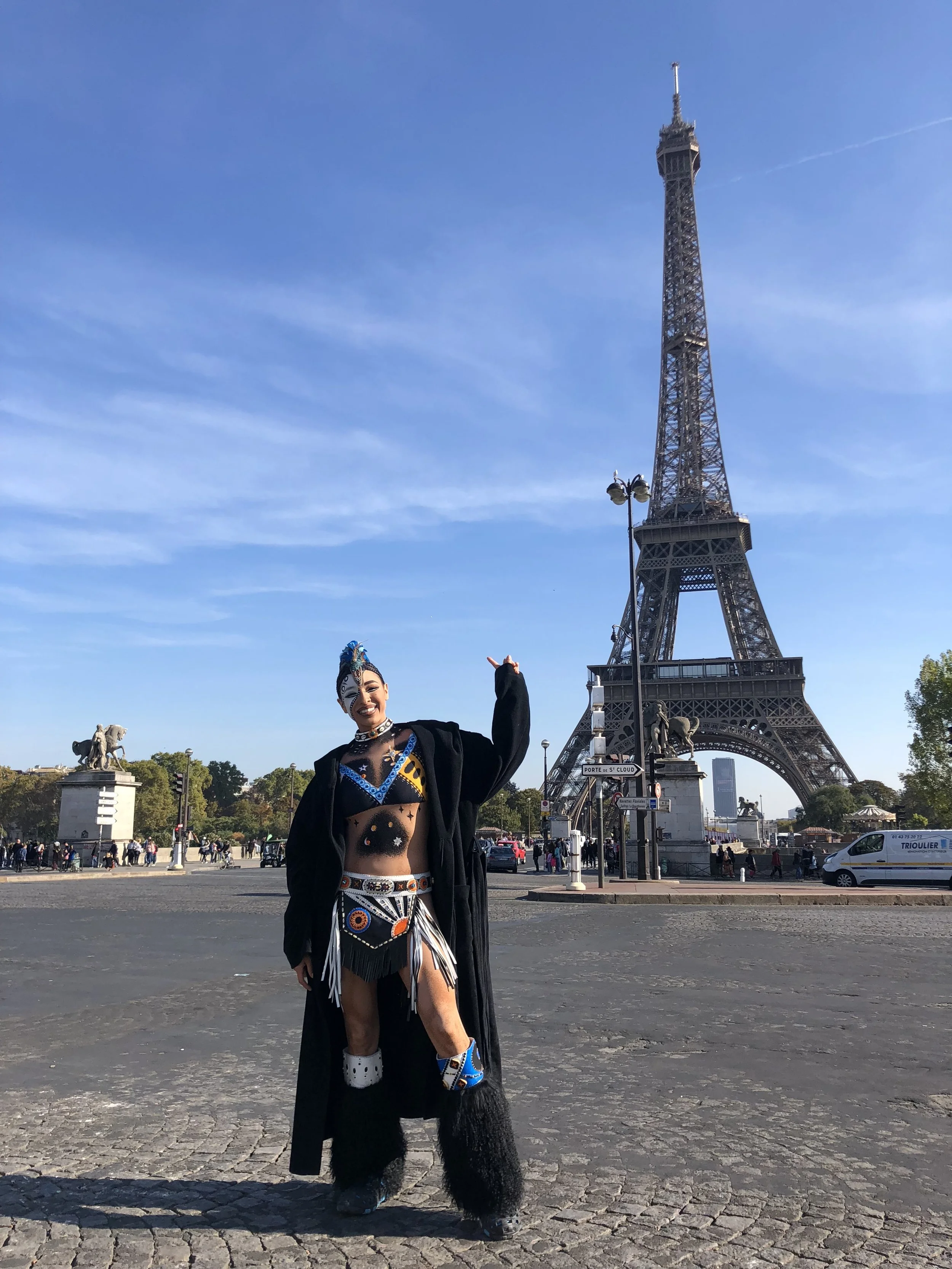 A woman dressed in a traditional indigenous costume standing in front of the Eiffel Tower in Paris, France.