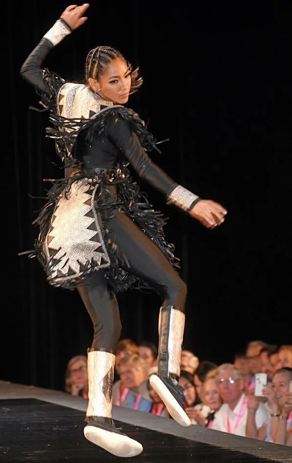 A woman performing a dance move on stage, wearing a black and metallic silver costume with fringes, black leggings with metallic accents, and oversized white shoes, with an audience watching in the background.