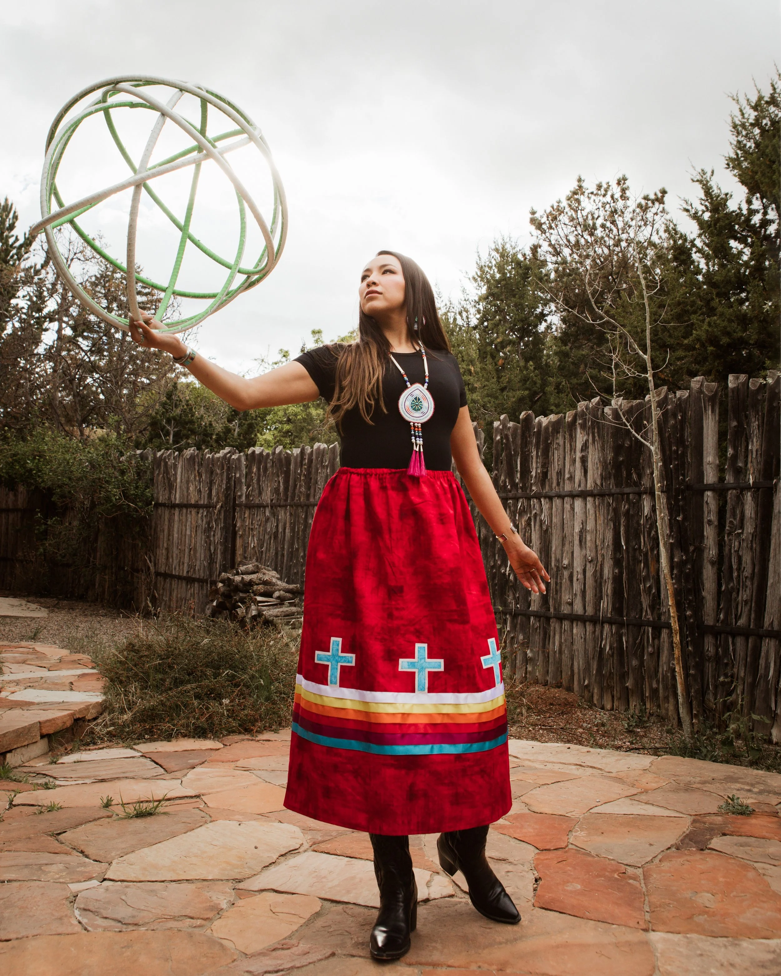 Shandien dressed in a black top and Native American ribbon skirt with colorful stripes and crosses, holding a large sphere made of hoops, standing outdoors on a stone path against a rustic wooden fence. Location Santa Fe, New Mexico.