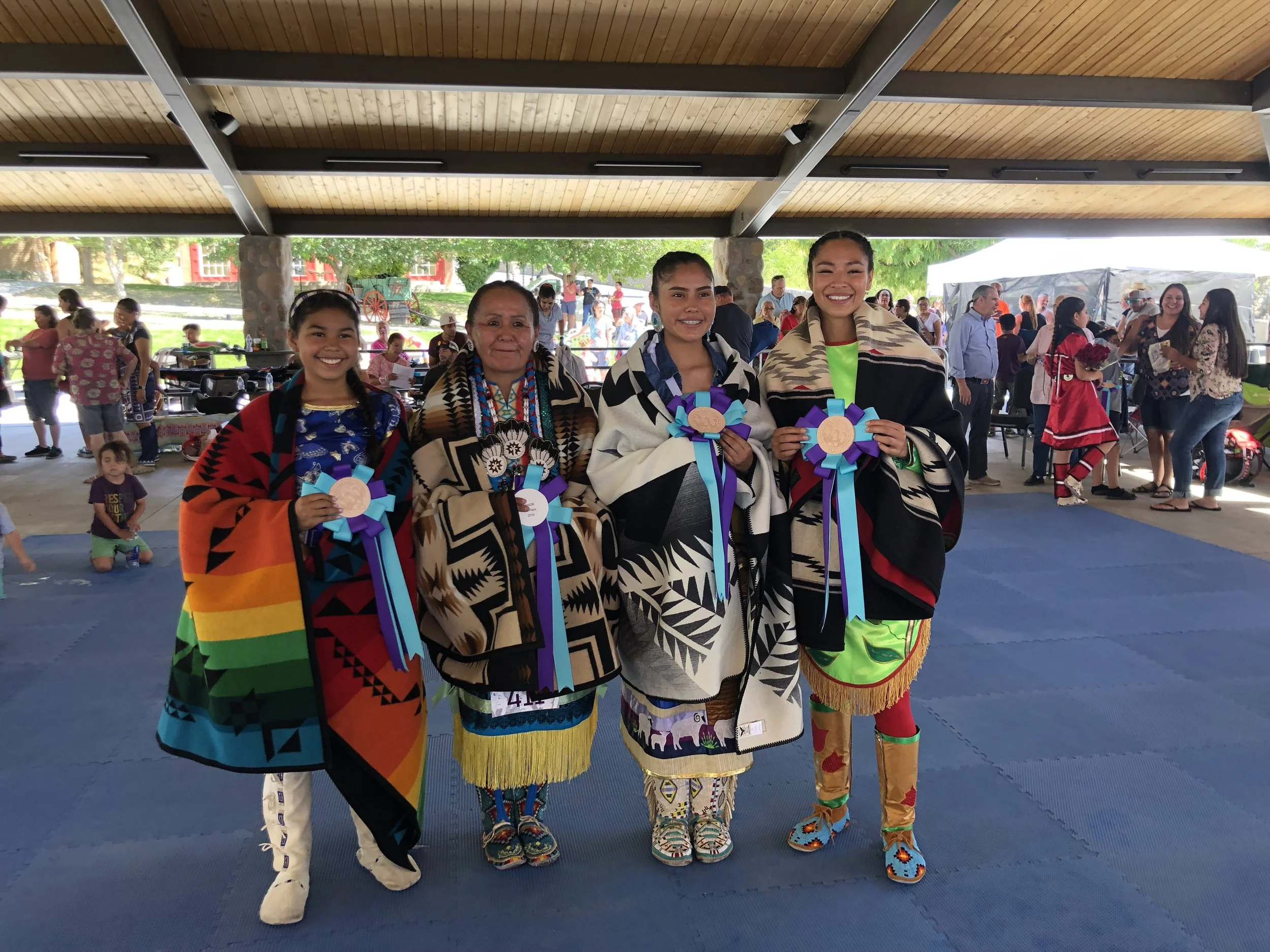 Four Indigenous women standing together at a cultural event, holding colorful ribbon awards, wearing traditional clothing and blankets, with a crowd of people in the background.
