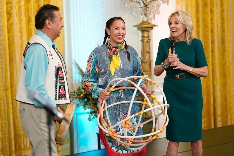 Three women on a stage, with yellow curtains and floral arrangements in the background. One woman in the center holds a colorful spherical object, and all three are smiling and engaging in conversation.