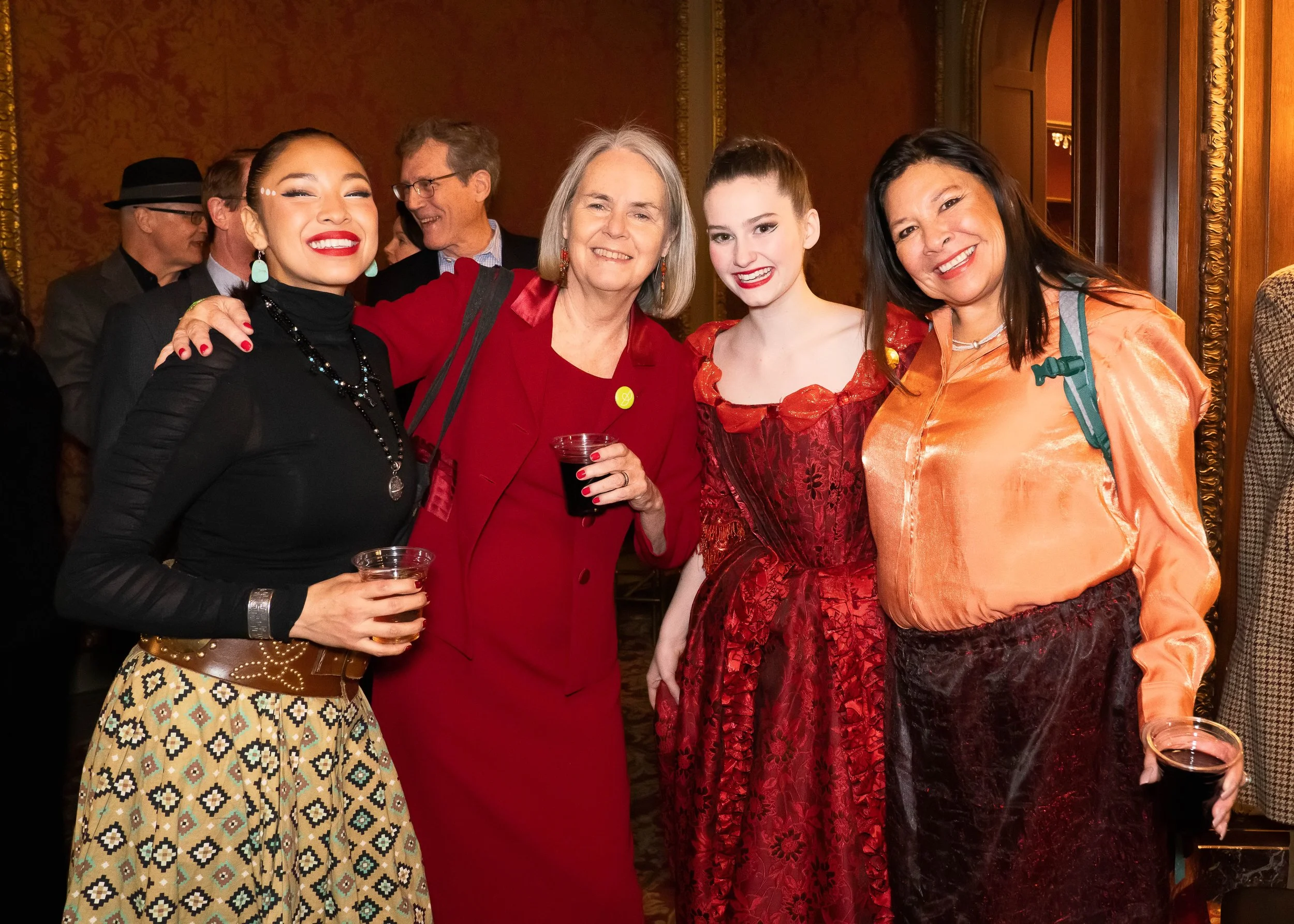 Group of five women smiling and posing together at an indoor event, dressed in colorful and stylish outfits, holding drinks.