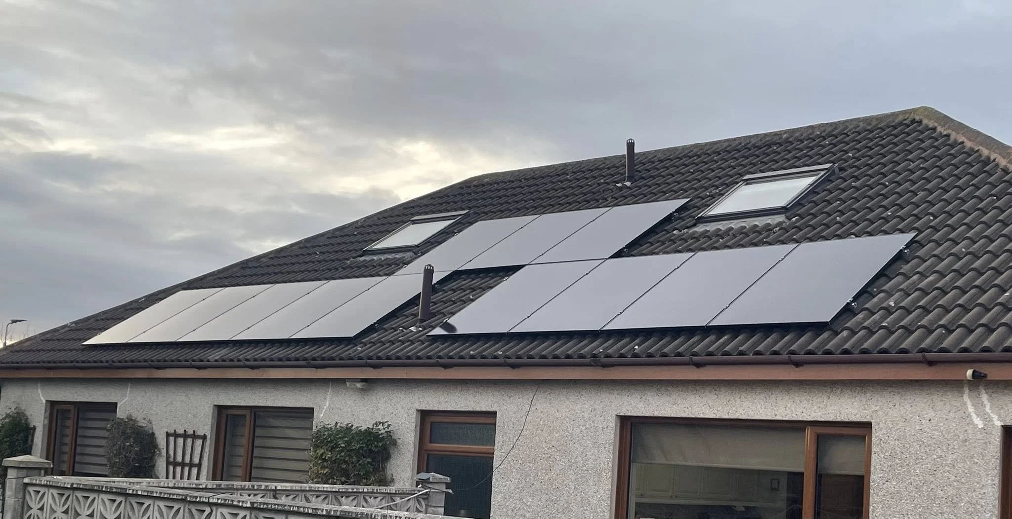 A house with solar panels installed on the roof and two skylights, against an overcast sky.