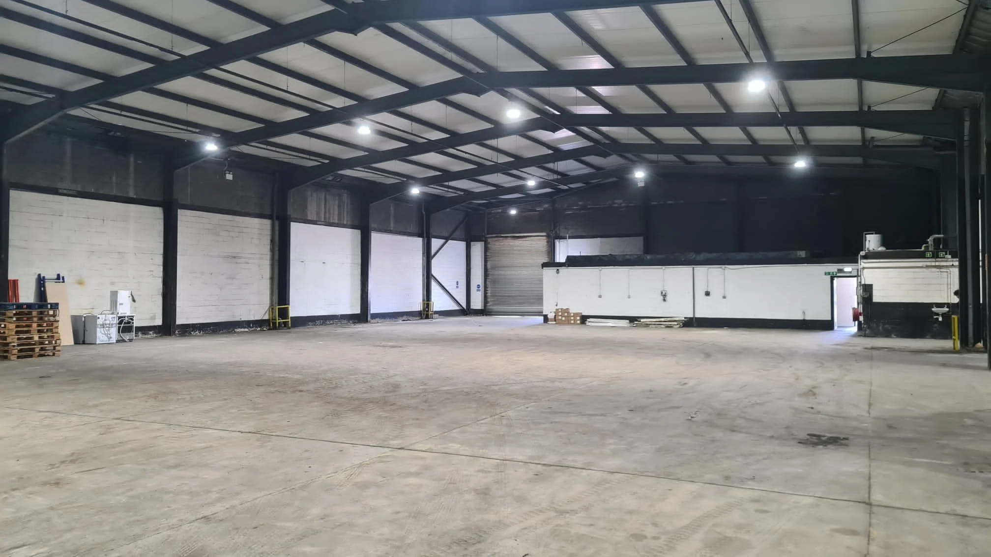 Empty industrial warehouse with high ceiling, exposed beams, bright overhead lighting, concrete floor, and minimal equipment along the walls.