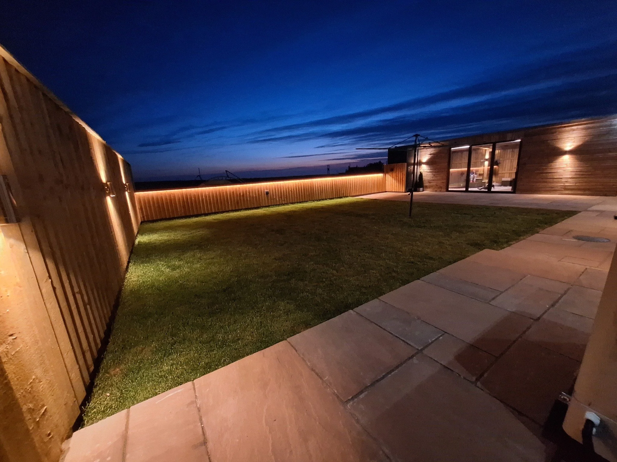 Nighttime view of a backyard with a grassy lawn, stone pathway, and a wooden fence with integrated lighting. A modern wooden house with large glass doors is visible on the right, with outdoor lights illuminating the space.